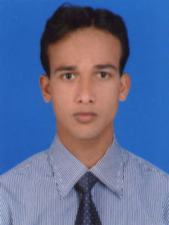 The image is a passport-style photograph of a person. The individual appears to be a young adult male with dark hair neatly combed to the side and styled in a modest manner. He is wearing a collared shirt with thin vertical stripes, presumably indicative of a formal dress code. The shirt has a dark color, which could be black or navy, and is paired with a dark blue tie featuring a pattern of light blue dots. The person's expression is neutral, and he has a clean profile, likely indicative of a personal portrait for identification purposes, such as a passport or ID. The background is a plain, solid blue, which provides a clear contrast to the subject, enhancing the visibility of the person's features.