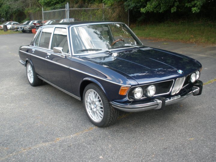 A black and white photo of a car parked in a parking lot - Pistonheads - The image features a classic blue BMW 2002 parked in a lot. It has two doors, and the multicolored taillights and side indicator lights are prominently visible. The car's overall condition appears to be good, with a polished finish and well-maintained paint. In the background, the setting suggests a quiet, possibly suburban, environment with some trees and parked cars.