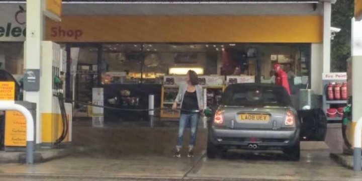 The BAD PARKING thread [vol3] - Page 197 - General Gassing - PistonHeads - This image is a photograph taken at an outdoor gas station. The scene captures a moment where a woman is standing next to a small silver hatchback car, seemingly engaging in a transaction. In the background, other customers and the interior of the gas station are visible, with various goods and services available. The gas station itself has a white and yellow combination, and a sign that reads "SHOP" extending above the station. The sky above appears overcast.