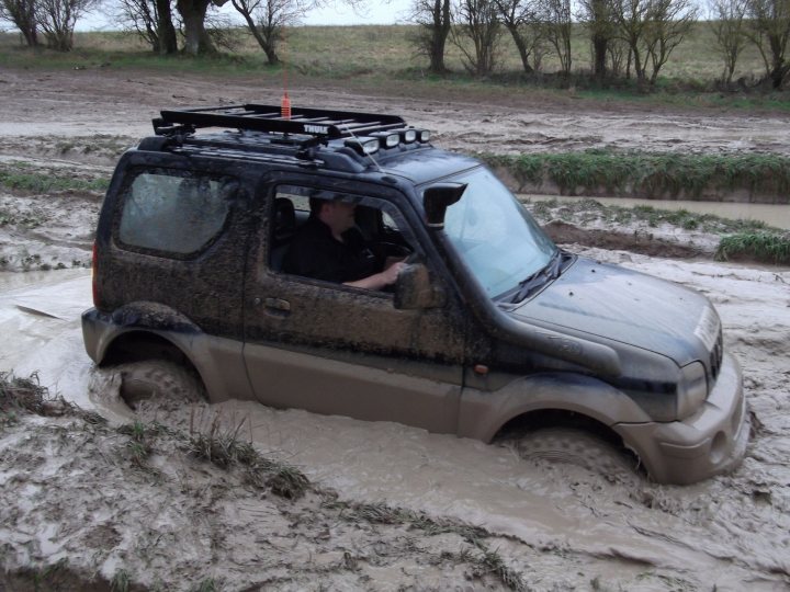 Pistonheads - The image showcases a challenging scene from off-road play. A black SUV is submerged in a muddy puddle, both the driver side and the passenger side, which is typical for vehicles attempting to traverse difficult terrain. The license plate on the back indicates the vehicle is registered in Arizona, although the registration details are not clearly visible. The tires are crowded in the mud, implying the driver may be attempting to reverse out of the situation. The environment around the puddle is overgrown, suggesting a pasture or a less-frequented dirt road.