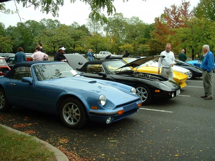 Can you import a TVR  S series into the US? - Page 1 - TVR in USA - PistonHeads - The image showcases a rich gathering of classic and sports cars parked in a lot. The most striking is the bright blue sports car, coupled with black. The cars are meticulously arranged against a backdrop of a green lawn and a mix of deciduous trees. A noticeable variety in the cars suggests a car show or a gathering of car enthusiasts. Indicative of a casual setting is the leisurely posture of the people standing around the cars.