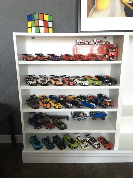 A display case filled with lots of different colored vases - The image displays a room with a white wall. On the left side of the frame, there is a large shelf that holds an assortment of toy cars. There are various colors and styles represented in this collection. To the right of the shelf, against the white wall, hangs a cube-shaped object resembling a Rubik's Cube. In the center of the image, partially obscured by the objects on either side, is an out-of-focus figure of a person. The overall setting suggests a playful or recreational space.