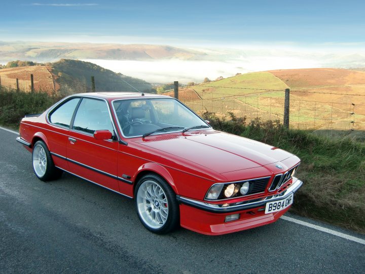 Eye pleasing cars - Page 6 - General Gassing - PistonHeads - The image shows a red BMW 5-Series vehicle on a winding road. The car appears to be a base model as indicated by the lack of visible V8 engine details or competitive badges. The setting is picturesque with a medieval mountain town in the background and a wooden fence running alongside the road. The sky is partially cloudy, suggesting a captivating scenic drive. The lighting on the car's side indicates it is likely to be late afternoon or early evening, casting a soft, golden glow on the scene.