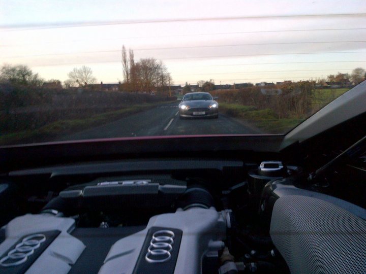 great day driving in yorkshire - Page 1 - Aston Martin - PistonHeads - The image is taken from the perspective of an Audi car's passenger seat, showcasing a scenic, winding road that appears to be entering a tunnel. The view captures a silver sedan driving ahead with its headlights on despite the daylight, indicating dawn or dusk. On the dashboard of the Audi, the engine bay with the car's emblem is visible, indicating the photo was taken inside the car from a driver's side. The environment outside the car suggests a rural or semi-urban setting with trees lining the road.