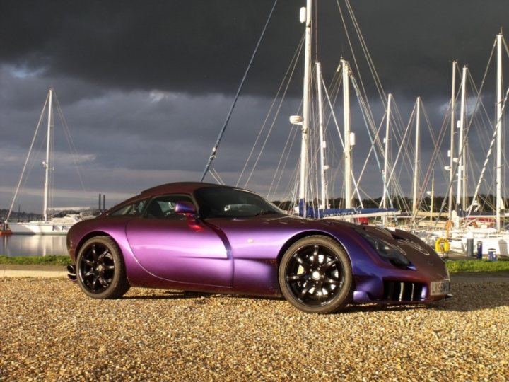 Moody Pistonheads Motor - This image showcases a **purple supercar** parked on a gravel surface. The car, viewed from the side, is sleek and has large, black rims. It is positioned in front of a row of sailboats docked in the background. The sky above is overcast, hinting at a possible change in weather conditions, and the grass beside is a vibrant green. The contrast between the purple car and the natural and maritime elements in the background creates a striking visual.