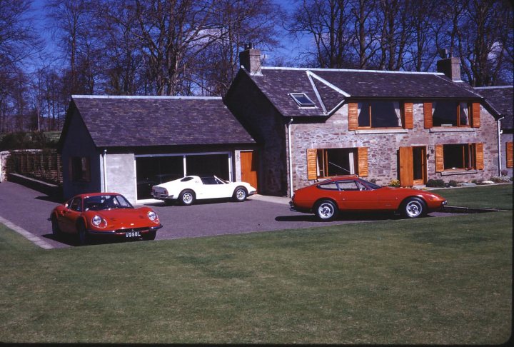 Pistonheads - The image presents a serene outdoor scene with a spacious lawn in front of two houses. The houses appear to be detached and are constructed with a stone-like facade. Parked in the driveway is an array of classic and vintage cars, varying in color from red to white, and from a car branded "Corvette." The vehicles showcase automotive design spanning different decades, adding to the overall vintage feel of the scene.