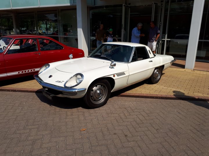 COOL CLASSIC CAR SPOTTERS POST!!! Vol 2 - Page 417 - Classic Cars and Yesterday's Heroes - PistonHeads - The image shows a vintage car on a brick pavement. The car is white and has a round headlight style. Various people are standing in the background, some near a red car. The setting appears to be a public area, possibly a parking lot or a park, in front of a building with large glass doors. The car takes up a significant portion of the frame, indicating its importance in the scene.