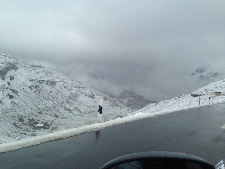 7 Countries 4 days & a banger - 'Help For Heroes' - Page 1 - South West - PistonHeads - The image shows a stretch of a snow-covered road leading into a cloudy valley. There's a large, dark mountain with snow-capped slopes in the background. The sky is overcast with a heavy deposit of snow, obscuring the details of the landscape. The road is untouched and currently not accessible due to the snow accumulation. The image is taken from the viewpoint of a vehicle, where the top of the steering wheel and a pair of sunglasses are visible in the lower right corner.