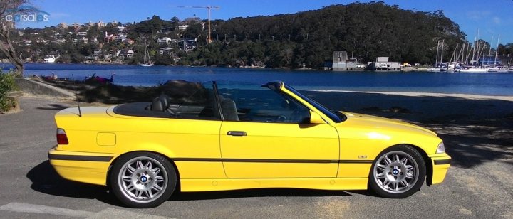 Daily Dreaming - what are you looking at? - Page 23 - Australia - PistonHeads - The image features a bright yellow convertible car parked on a paved lot. The car is a two-door model, indicating that it provides space for two passengers, with seating facing each other. The car is positioned facing towards the right side of the image, with its top down, allowing the interior to be seen, including the two front seats. The setting appears to be outdoors, as there is an open blue sky in the background, suggesting a pleasant day. In the distance, you can see a serene body of water, possibly a river or a lake, which contrasts the urban setting with the natural landscape. A few boats are visible on the water, adding to the recreational atmosphere of the scene.