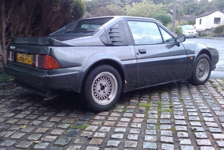 G32 - Page 1 - Ginetta - PistonHeads - The image features a small gray car parked on a cobblestone street. The car is an older model, with a single visible headlight and a noticeable spoiler at the back. It has a license plate starting with the letters "K50." The vehicle appears to be of European origin, possibly a UK model given the plate format. The car stands out against the textured backdrop consisting of a wall trimed with plants and a driveway.