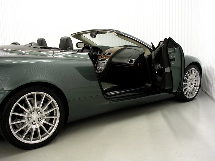 The non-black-non-grey-non-silver thread - Page 19 - Aston Martin - PistonHeads - The image shows a sleek, two-door sports car parked indoors on a light grey floor with textured wallpaper in the background. The vehicle is a convertible with a black interior, a whitewalled wheel, and a prominent side gill. The sideview mirror is designed with a high-mounting style, which is typical for sporty vehicles. The car's color scheme includes a body hue that combines black with metallic green or silver elements.