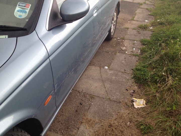 A car parked next to a parking meter - Pistonheads - The image features a grey sedan parked on the side of a road. A small piece of litter, possibly a food wrapper, is visible beside one of the car tires. There is a broken windshield on the driver's side, resulting in visible damage. The vehicle is parked on a grassy area with cracked pavement or possibly a sidewalk adjacent to the grass. It appears to be daytime, and no people or other cars are visible in the immediate vicinity of the car.