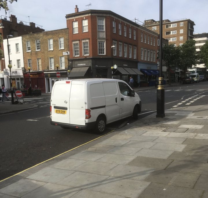 The BAD PARKING thread [vol4] - Page 517 - General Gassing - PistonHeads UK - The image depicts a quiet city street scene. A white van is parked on the side of the road, adjacent to a curb and near some buildings. There's another vehicle partially visible in the distance, suggesting this might be a busy area despite the calm appearance. The street has a few shops or businesses along it, which adds to the urban setting. The sky appears overcast with no direct sunlight. There are no people or moving vehicles visible in the immediate vicinity of the van.