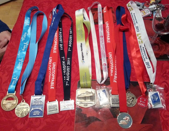 Displaying running medals - Page 1 - Sports - PistonHeads - The image shows a colorful collection of sports medals and ribbons displayed on a red surface. There are a total of nine medals, each with different designs, colors, and ribbons. They are arranged in a curve, with some medals hanging on red ribbons and others tied to silver pins. The background reveals a red tablecloth and a person's leg visible at the left edge of the frame, suggesting that this is a home environment. The overall presentation suggests these medals have been earned and are being celebrated or displayed with pride.