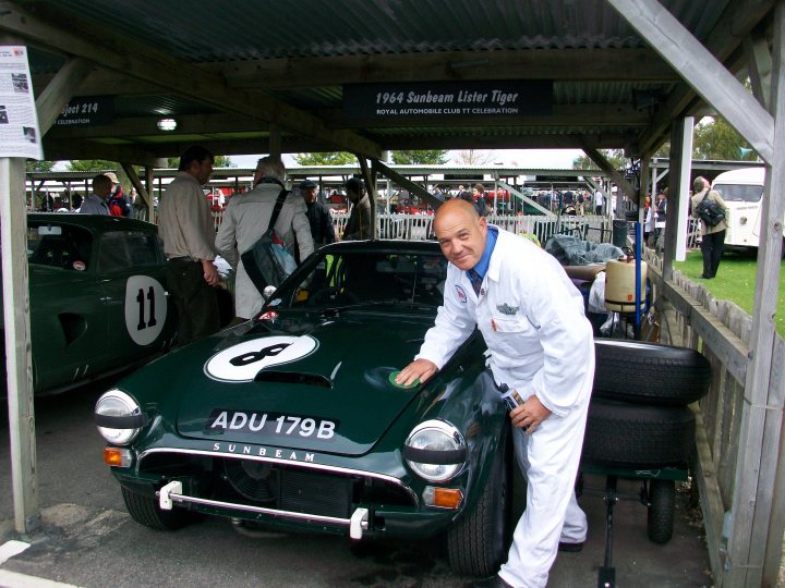 Pistonheads - The image depicts an older man dressed in a suit, standing proudly next to a green sports car. The car is adorned with a variety of advertisements and sponsors, indicating its likely participation in a racing event or competition. The setting appears to be a car dealership or exhibition area, as suggested by the presence of other cars and various people in the background. Notably, the car's design and the man's attire evoke a sense of nostalgia and elegance. The man seems to be posing for the photo, possibly acknowledging his role in the car's history or performance.