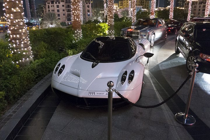 Middle East spotted thread - Page 63 - Middle East - PistonHeads - The image depicts an exotic white sports car with headlights and taillights on, parked at a valet station during nighttime or twilight. The car is an electric vehicle, as indicated by the charging connector on the front. It appears to be a high-end model, given its sleek design and clean lines. A valet attendant or rope is managing access to the car, as indicated by the roped-off area preventing direct approach. In the background, there are additional vehicles, suggesting that the setting could be a social gathering or event.