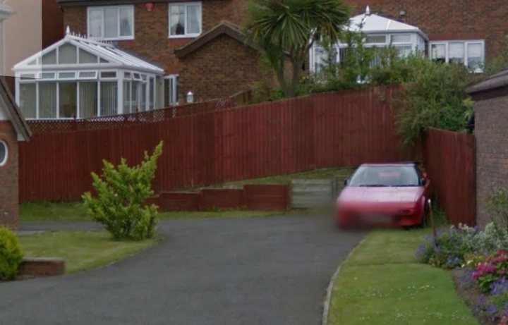 Spotted Ordinary Abandoned Vehicles - Page 49 - General Gassing - PistonHeads - The image shows a residential area with a variety of buildings. In the foreground, there is a driveway leading to a red car parked on the right. The car is captured in motion, with its license plate clearly visible. Behind the car, there is a section of a wooden fence that divides the driveway from the public road. A well-maintained garden with green lawn and various flowers is noticeable on the left side of the image. The buildings in the background appear to be brick houses with several windows, and one of them has a white canopy.