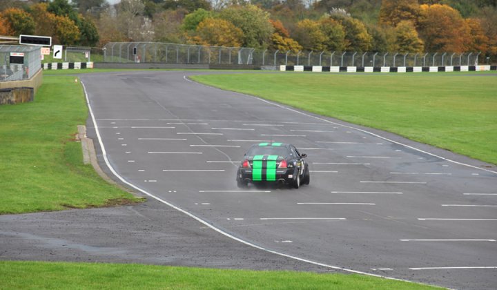 Pistonheads - The image is a vibrant daytime photograph capturing the exterior of a car racing on a winding paved race track. The car has a body wrap design in green and black stripes and a large rear wing. The track is nestled amidst lush green and autumn-colored foliage, with spectator grandstands visible on the right side of the track. Safety barriers can be seen at intervals along the track, suggesting that the environment is controlled for competition purposes. The composition of the image highlights the motion of the car, as indicated by the blurred wheels, in contrast to the stark, clear presence of the car and track markings.