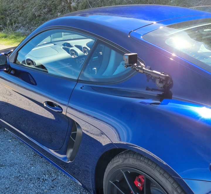 Pistonheads - The image shows a close-up of the side view of a sports car. It has a vibrant blue color and appears to be a high-performance model, judging by its sleek design and aerodynamic features. Mounted on the side mirror is a camera that seems to be recording or monitoring the vehicle, possibly for security purposes. The car is parked in what looks like an outdoor setting with trees and a road visible in the background.