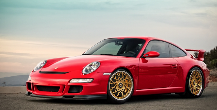 A red car is parked on the side of the road - Pistonheads - The image showcases a vibrant red Porsche 911 GT2 RS parked on a paved surface, capturing the side profile view of the car. The GT2 RS is known for its unique color scheme with gold accents, and this one features a matching gold brake caliper. Its elongated hood reveals the powerful engine, and the aggressive front bumper and side skirt hint at its sportscar character. The background suggests an outdoor setting, possibly during a sunset, with a hint of the sky and the silhouette of trees.