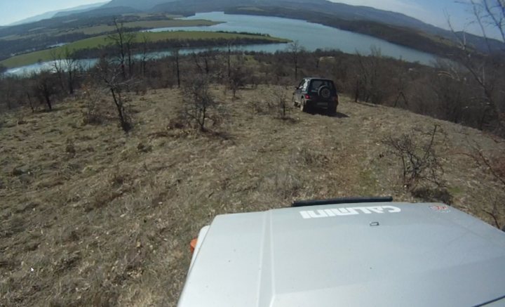 Bit of off-roading, and a bit of camping. - Page 1 - Off Road - PistonHeads - The image captures a breathtaking panorama from a high vantage point overlooking a coastline. The viewer is positioned in a rugged, rocky terrain with sparse vegetation, seemingly in the middle of an adventure. Below, there's a serene body of water with a boat cruising on it, offering a sense of tranquility. In front of the viewer is an SUV, indicating that they are possibly at the end of their journey, standing on the edge of a cliff or a hill. The sky above is partially cloudy, casting a soft light over the entire scene.