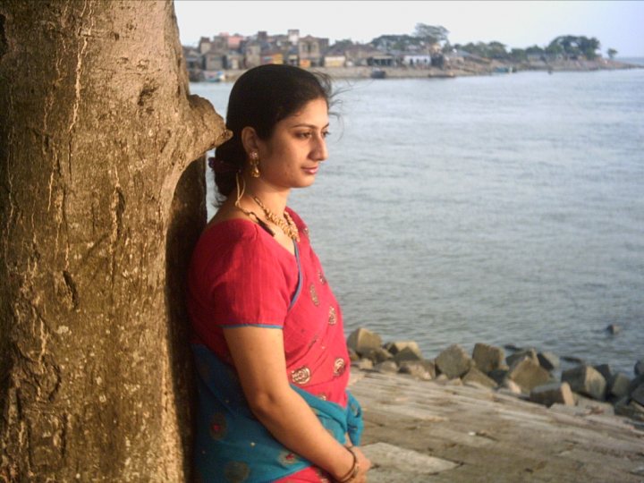 In the image, a woman with long hair is seen standing, leaning slightly back against a large tree by the water. She is wearing a red traditional outfit with a colorful blouse and skirt. The woman appears relaxed, looking into the distance over the water. The background reveals a tranquil scene with a calm body of water, a distant shore with a few buildings, and a clear sky overhead.