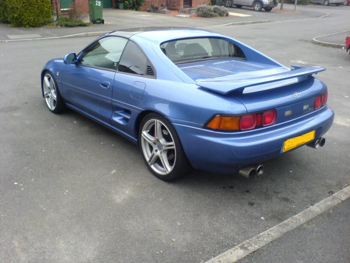 Pistonheads Write Shes - The image captures a scene on a street where a vintage blue sports car is parked. The car features a sunroof and a visually appealing design with a long rear boot and a distinctive spoiler. The tail lights are bright, and the car is equipped with gold trim, which adds a touch of elegance. The car's license plate is prominently visible on the back. In the background, there are other parked cars and a green trash can. The street appears to be in a residential area, and there are no people visible in the image.