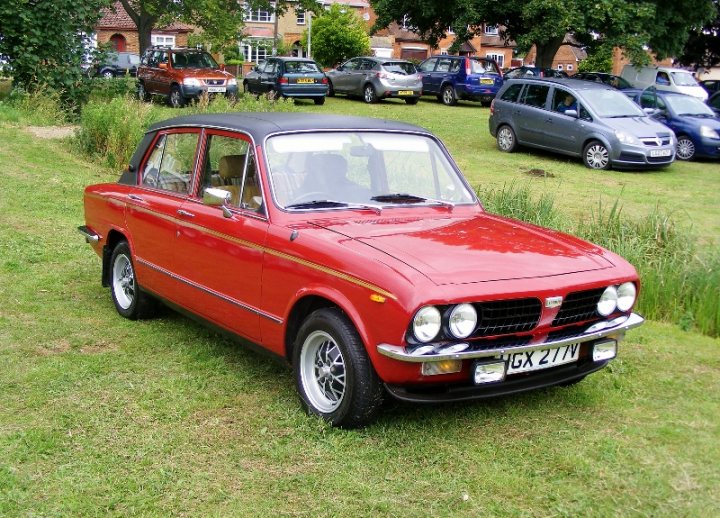 COOL CLASSIC CAR SPOTTERS POST!!! Vol 2 - Page 59 - Classic Cars and Yesterday's Heroes - PistonHeads - The image shows a red vintage car parked on a grassy area. The car is surrounded by a variety of other cars and parking lots, which provide a contrast in color and style. The surroundings include a chain-link fence and it's not clear where the car is located without additional context. The vintage car is the main focus, capturing attention with its bright color and classic design.