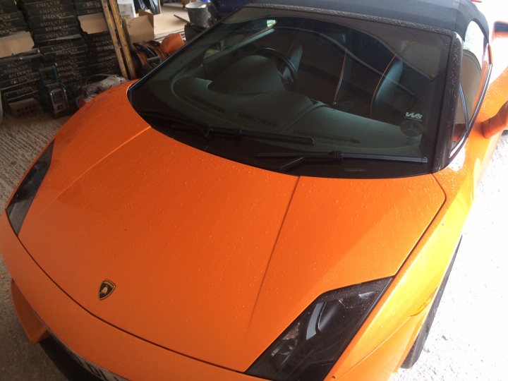 More pics of my LP560 - Page 1 - Gallardo/Huracan - PistonHeads - The image is a photograph showcasing an orange sports car with textured paint. The car appears to be parked indoors, possibly in a warehouse or storage area, as suggested by the surroundings that include shelving in the backdrop. The hood of the car is slightly ajar, revealing the engine area underneath. The car is depicted in profile view, highlighting its low, sloping platform and distinctive design.