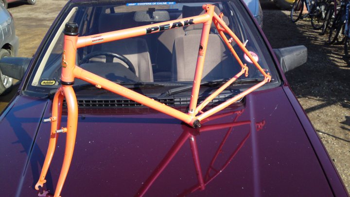 Recumbent Dual Suspension Pistonheads - This image features an abandoned bicycle on the ground in front of a car. The bicycle has an orange frame and black and white accents, while the car appears to be parked on a gravel surface with a blue-grey hue visible through its windows. The bicycle's front wheel is closer to the camera, while the back wheel is further away and more obscured. There's a slight reflection of the bicycle on the car's shiny surface.