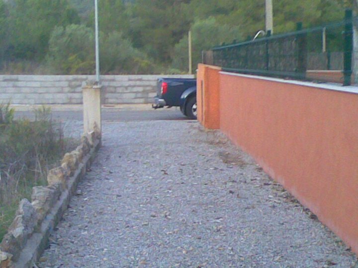 Pistonheads - The image shows an outdoor scene, focusing on a path bordered by a rock wall on one side and a vibrant red wall and orange fence on the other. To the right of the path, there's a mowed area with bare ground and a few areas of green grass and vegetation. A black pickup truck is parked on the road beside the path, facing away from the viewer. The truck is mostly visible from the side. The ground in the foreground appears to be made of rock and gravel, leading up to the path. The overall scene suggests a maintained area, likely a residential or public park.