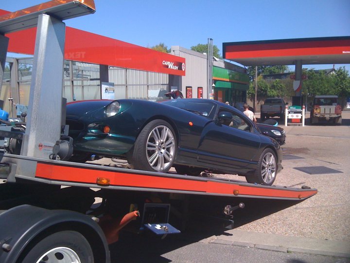 Ive Elite Pistonheads Joined - This image features a dark-colored, low-slung sports car perched on the bed of a tow truck. The car is facing away from the viewer, revealing its sleek design and shiny exterior. The tow truck is positioned on a road or parking lot, as indicated by the presence of other vehicles in the background; some vehicles are parked while others are moving. The setting suggests this might be a roadside assistance scenario or a car dealership.