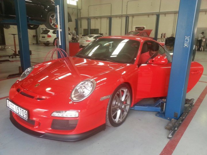 A red car is parked in a parking lot - Pistonheads - The image features a vehicle lift in a workshop, with a man standing next to a red sports car. The car is suspended in the air by the lift, which has a red, white, and blue striped pole to secure it. The interior of the car appears to be in the process of being modified or inspected, possibly being fitted with racing seats. Another car can be seen in the background undergoing similar maintenance or inspection. The scene suggests a setting focused on upgrading or servicing sports cars.