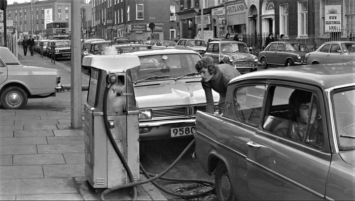 A 'period' classics pictures thread (Mk III) - Page 33 - Classic Cars and Yesterday's Heroes - PistonHeads UK - The image is a black and white photograph showing an urban street scene. There are several cars parked along the side of the road, with one car prominently featured in the foreground. People can be seen walking on the sidewalks. The vehicles are older models, indicating that this photo might have been taken some time ago. In the background, there is a gas station visible, suggesting that this is a busy commercial area.