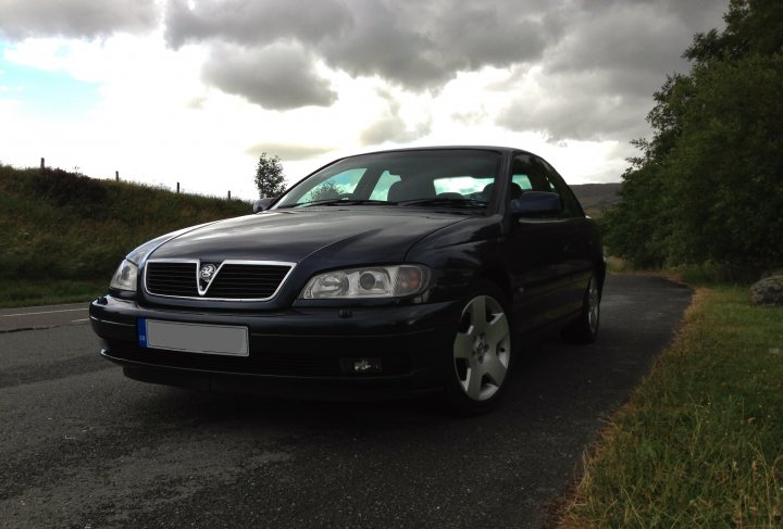 2002 Omega 3.2 V6 Elite Saloon - Page 1 - VX - PistonHeads - The image depicts a dark-colored sedan parked by the side of a road. The car's sleek design suggests it could be a model from a luxury brand. The setting appears to be at dusk, as suggested by the dim lighting and the approaching darkness. The road is lined with lush green trees and a fence can be seen against the sky. A lone cloud looms in the background, adding a dramatic effect to the scene.