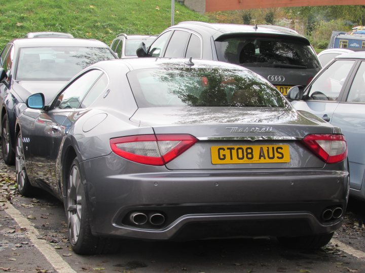 Supercars spotted, some rarities (vol 6) - Page 469 - General Gassing - PistonHeads - The image portrays a line of cars parked in a lot, with the front of a sports car in the foreground. The make of the car is clearly visible, as it reads "GT08 AUS." Another brand name can be discerned, but it is not a luxury or high-end vehicle. The setting appears to be an outdoor parking area, possibly next to a garden, as there are plants in part of the background. A few other parked cars can be seen in the background, though their makes and models are not identifiable.