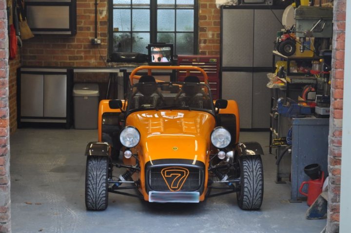 Who has the best Garage on Pistonheads???? - Page 41 - General Gassing - PistonHeads - The image depicts a workshop setting inside a garage. The main focus is a vibrant orange Bugatti car parked in the center. It features two-passenger seating and a small inspection window in the front. The car is designed with a hood scoop and a driver's seat on the left side of the vehicle. The garage is equipped with various tools and equipment, indicating a space for repair or modification of vehicles. The walls of the garage show a brick construction and are embellished with a few pictures, which add a personal touch to the space.