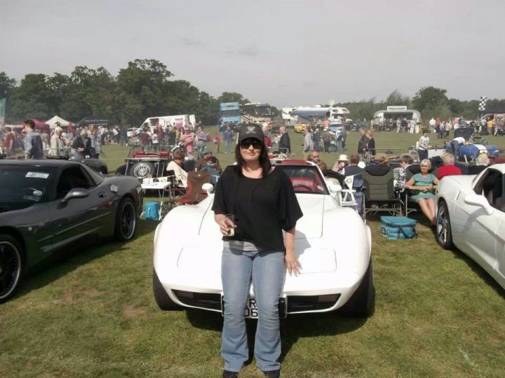 Pistonheads - The image shows a bustling street scene with a crowd of people mingling around and a variety of vehicles parked, including cars and a motorcycle. In the center, a woman is standing, appearing to be the focal point of the image. The setting appears to be an outdoor event, as indicated by the presence of numerous chairs and tents. The environment suggests a social gathering with leisure vehicles and attendees.