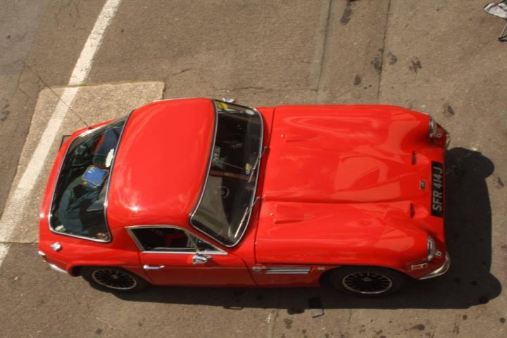 Early TVR Pictures - Page 90 - Classics - PistonHeads - The image showcases a vibrant red vintage car, rich in color and shiny in appearance, parked on the side of a road. It has a white stripe on the side, adding to its appeal. The car is the focal point of the image, set against a backdrop of a plain concrete surface and a yellow curb. Despite the simplicity of the background, the red and white color scheme stands out, creating a striking contrast. The car's well-maintained condition and neat parking suggest a sense of pride and ownership.