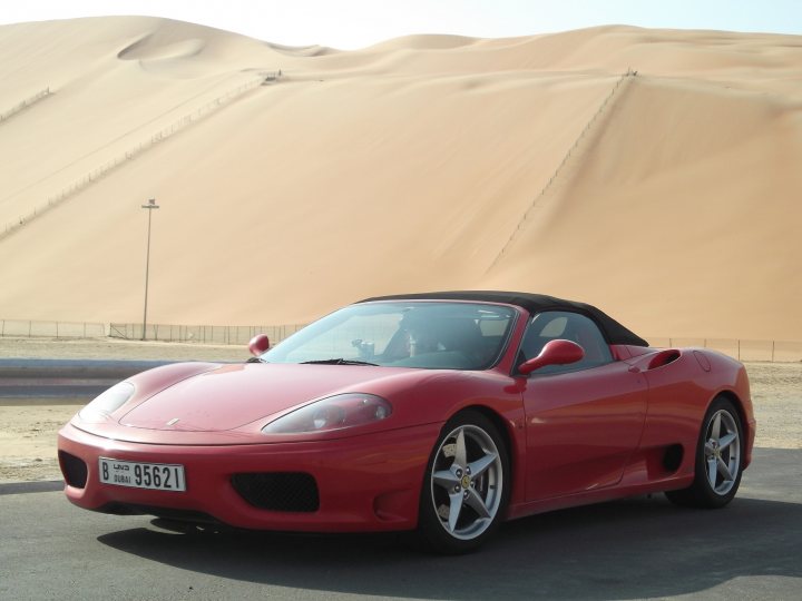 A car with a surfboard strapped to the back - Pistonheads - The image showcases a striking red Ferrari parked on a sandy patch of a sand dune desert, with noticeable tire tracks. The car is positioned in the lower left corner of the frame, drawing attention to its two-door, convertible design. The vast expanse of brown sand dunes forms the backdrop, emphasizing the solitude and freedom the scene conveys. The car appears to be abandoned or left unattended, adding a touch of mystery to the otherwise serene landscape.