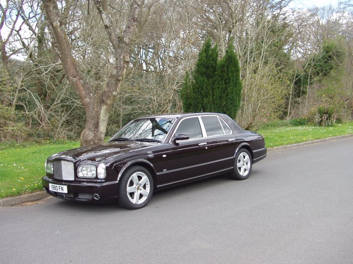 A car parked on the side of the road - Pistonheads - The image shows a dark-colored luxury sedan parked on a roadside. The car has a polished finish and is equipped with chrome grille and a sunroof. The car features multiple large wheels, which typically designates high-end vehicles. In the background, there are trees and a grassy area, suggesting a suburban or less populated residential district. The sky is overcast but does not detract from the parked vehicle.