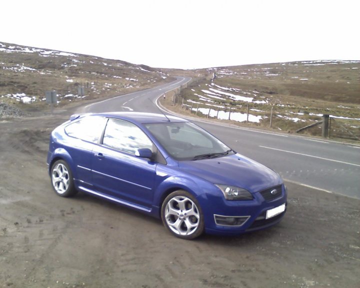 Good Places to Photograph your car - Page 1 - General Gassing - PistonHeads - The image showcases a blue hatchback parked on a gravel shoulded alongside a paved road. The car is set against a backdrop of a rural landscape, with a large hill and a fence along the roadside. The weather appears to be cloudy, suggesting a potentially cool day. The overall atmosphere of the image is calm and tranquil, capturing a quiet moment in what seems to be a serene setting.