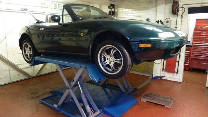 are MK1 and MK2 springs the same? - Page 1 - Mazda MX5/Eunos/Miata - PistonHeads - The image features a scene from a garage where a black two-door sports car is being serviced on a lift. The lift is supported by a sturdy blue frame, and the car is securely positioned on its black tires with silver rims. The car's suspension is clearly visible beneath the lift. In the background, tools and equipment are organized on shelves, contributing to the professional atmosphere of a well-equipped workshop. The garage itself has a tiled floor, suggesting a high degree of cleanliness and maintenance.