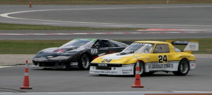 A car is parked on the side of the road - Pistonheads - The image is a color photograph that shows two race cars on a slightly raised racing track. The car in the foreground is predominantly yellow with black and red stripes, while the other car is predominantly black with white and red stripes. Both cars bear numbers and sponsor names, suggesting they are part of a racing competition. The setting appears to be a professional racing event. The background is out of focus, emphasizing the cars in the foreground.