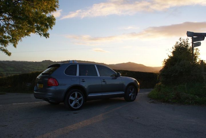 Touareg roof rack/spolier on a cayenne? - Page 1 - Porsche General - PistonHeads - The image captures a tranquil scene with a silver SUV parked on the side of a rural road. The car's modern lines and shiny surface contrast with the natural surroundings. A vibrant sunset casts a warm glow on the landscape, with light reflecting off the car's surface, suggesting the sun is low on the horizon. Beyond the car, the road stretches out into the distance, edged by lush green hills that roll gently into the horizon. To the side, a lone tree stands, its silhouette adding depth and texture to the scene.