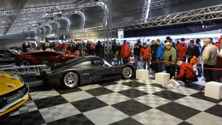 Grasserultima God Carbon Pistonheads - The image shows a bustling auto show, where a vintage car exhibit has drawn a crowd. The centerpiece of the image is a black sports car, displayed on a grey and black checkered floor, which resembles the classic motif of a racing starting line. The car is surrounded by enthusiastic attendees who have gathered around, some of whom are holding papers, possibly reviews or pamphlets. The backdrop of the exhibit features a stage with hanging lights, enhancing the atmosphere of the car show. The image captures the excitement and anticipation of attendees as they explore and appreciate the sports cars on display.