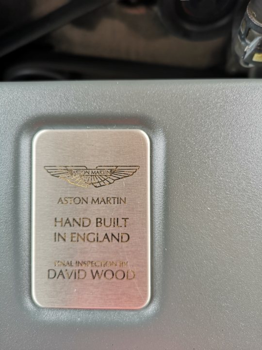 So who did your 'Final Inspection'? - Page 10 - Aston Martin - PistonHeads - The image features a metallic badge prominently displayed in the center. It appears to be a plaque or emblem with text that includes the names 'Aston Martin' and 'Handbuilt in England.' Additional text mentions 'JAMES BOND 007,' suggesting it is related to James Bond, known from the James Bond film series. Below this main text, there is a signature or name written 'DAVID WOOD,' indicating it may be an autograph or a special edition badge. The background is blurred but gives the impression of a mechanical or technical setting, hinting at the potential association with automotive engineering, which is consistent with Aston Martin's brand image.