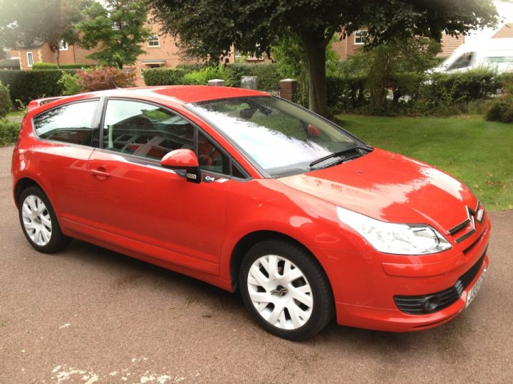 A red car is parked in a parking lot - Pistonheads - The image shows a red two-door car parked on a driveway. The car appears to be a hatchback model, possibly a compact or small family car, with clear headlights and a glossy finish. It's positioned in profile, highlighting the styling of the vehicle, including the wheels which are visible on the lower part of the image. The background indicates that the car is parked in a residential area, in front of a house, with trees and an expansive lawn visible.