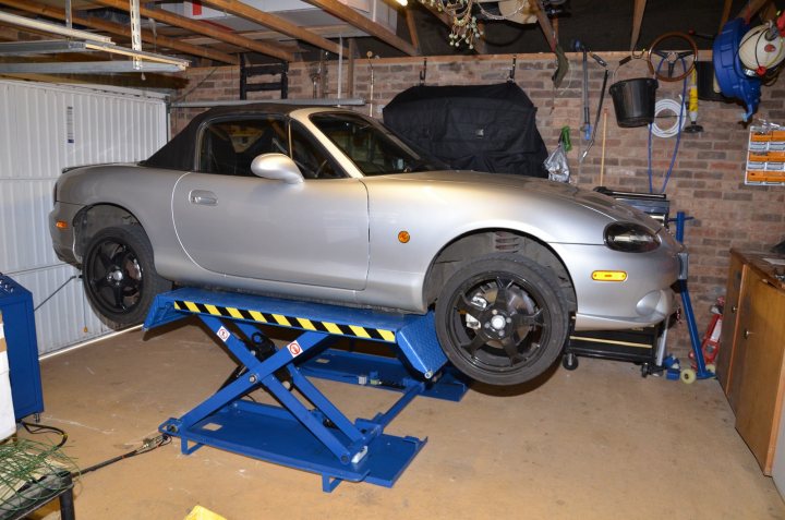 Garage Pistonheads Ramp Buying - The image shows a silver compact car sitting on a two-post car lift inside a garage. The vehicle is secured with safety straps and has been raised to a height consistent with the lift's design. The lift appears to be of a manual screw-down type, with the front two posts extending upwards and the car's wheels resting on corresponding guides. The garage surrounding the car lift has an uncluttered appearance, with no visible storage facilities in the immediate vicinity.