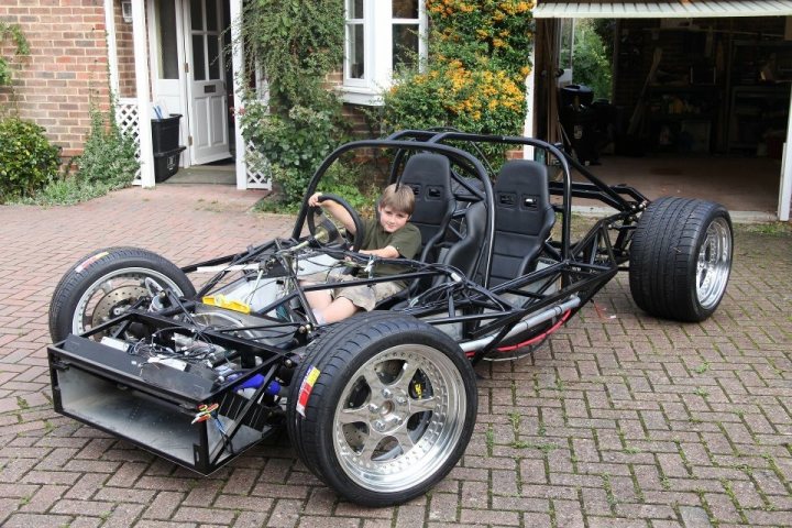 Good place to purchase unfinished ultimas? - Page 1 - Ultima - PistonHeads - The image shows a young boy sitting in the driver's seat of a large, black quad-wheeler or off-road vehicle. The vehicle is parked a few feet away from a residential house with a white door frame and a brick driveway. The boy appears to be alone inside the vehicle, and he has his arms on the steering wheel as if he is pretending to drive. The vehicle has multiple wheels, and there are metal supports visible around the driver's seat, likely for safety or structural support. The outside appears to be an English-style terrace, suggesting the vehicle is a unique toy car designed for play indoors.